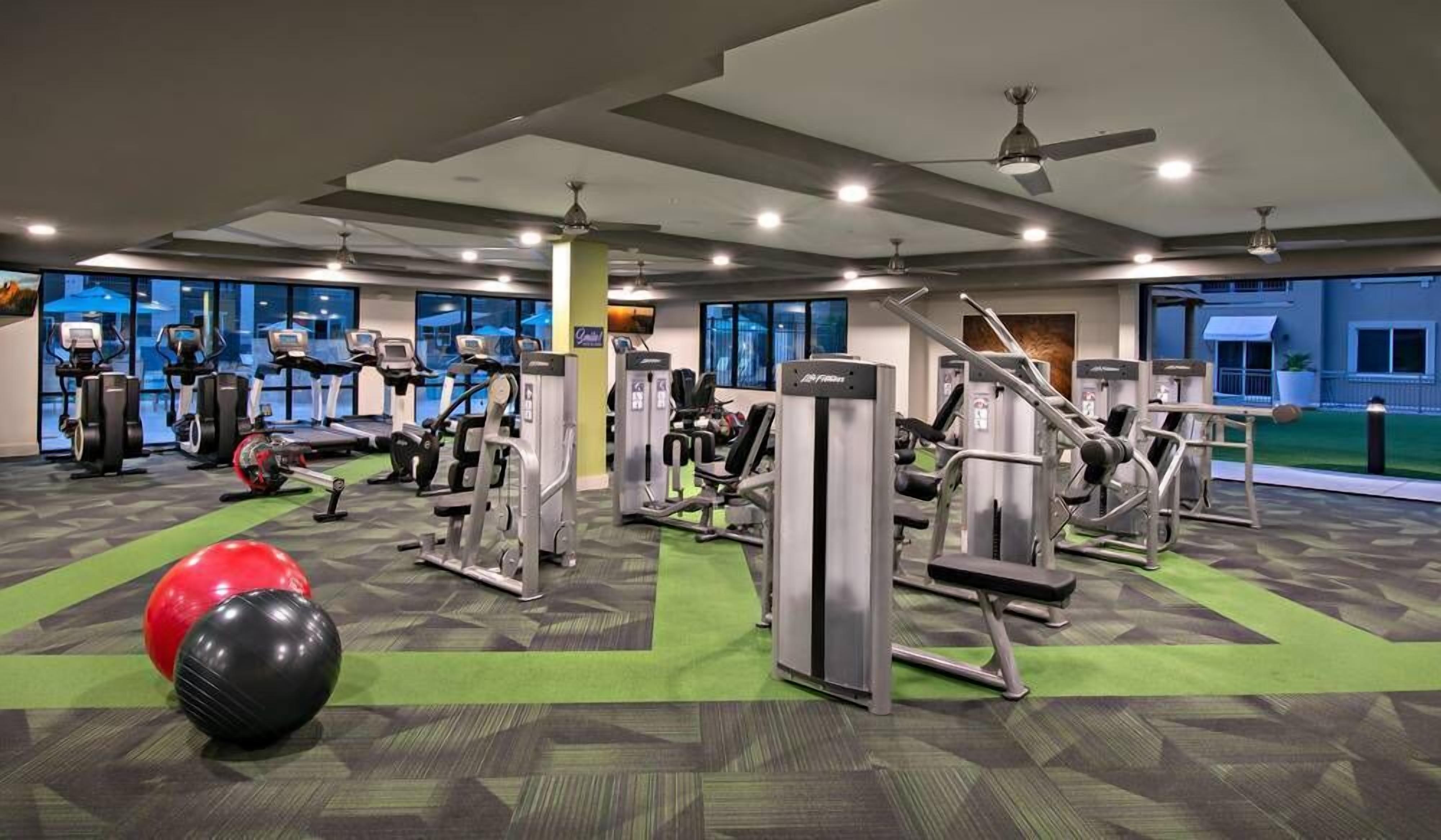 Ein Fitnessraum mit unterschiedlichen Trainingsgeräten, einem grünen Teppich und großen Fenstern.