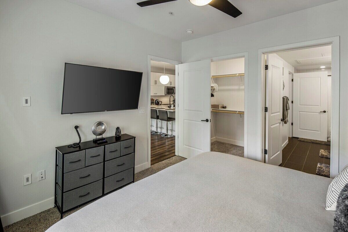 A bedroom with a bed, a TV mounted on the wall, a dresser, and an open door leading to a kitchen area.