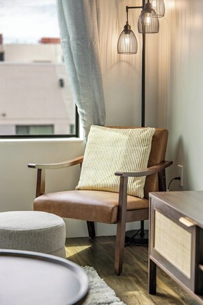 A modern chair with a cushioned backrest and wooden armrests, positioned next to a round ottoman and a small side table with a drawer. - Escape To The Tempe Penthouse King Bed Studio (Tempe)