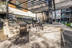 Eine Terrasse im Freien mit einer Grillgelegenheit aus Stein, Sitzmöbeln und Blick auf ein modernes Gebäude.
