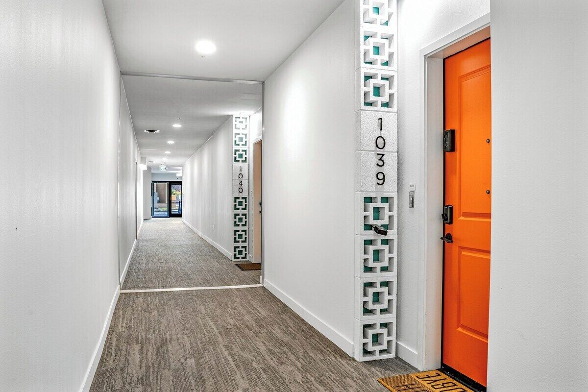 A hotel hallway with a carpeted floor, white walls, and a bright orange door.