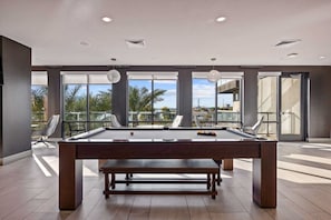 Une table de billard dans une pièce spacieuse, dotée de grandes fenêtres et offrant une vue sur des palmiers.