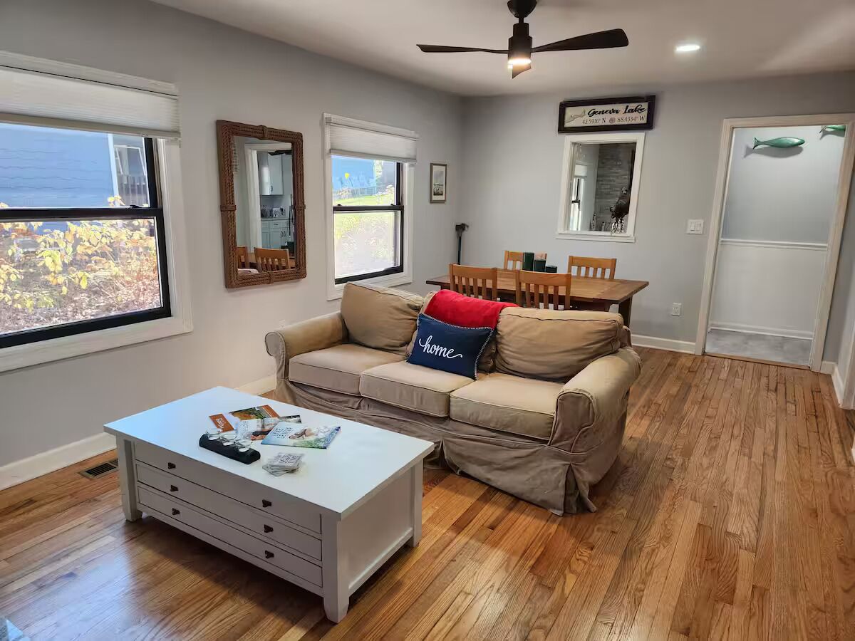 A living room with a beige sofa, a white coffee table, and a dining table with chairs. There are windows with blinds, a ceiling fan, and a door with a fish decoration.