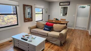 A living room with a beige sofa, a white coffee table, and a dining table with chairs. There are windows with blinds, a ceiling fan, and a door with a fish decoration.