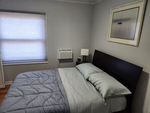 Une chambre à coucher avec un lit, une fenêtre avec des stores, un climatiseur fixé au mur, une table de chevet avec une lampe et un tableau encadré accroché au mur.