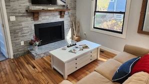A living room with a fireplace, a television mounted on a stone wall, a window with a view, and a beige sofa.