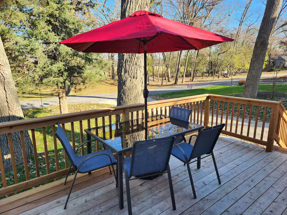 A wooden deck with a table and chairs, a red umbrella, and a view of trees and a road.