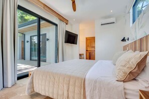 A bedroom with a large bed, a television mounted on the wall, and a sliding glass door leading to an outdoor area. - Casa VistaMar Spacious 5-bedroom villa built around a tree! (Potrero)