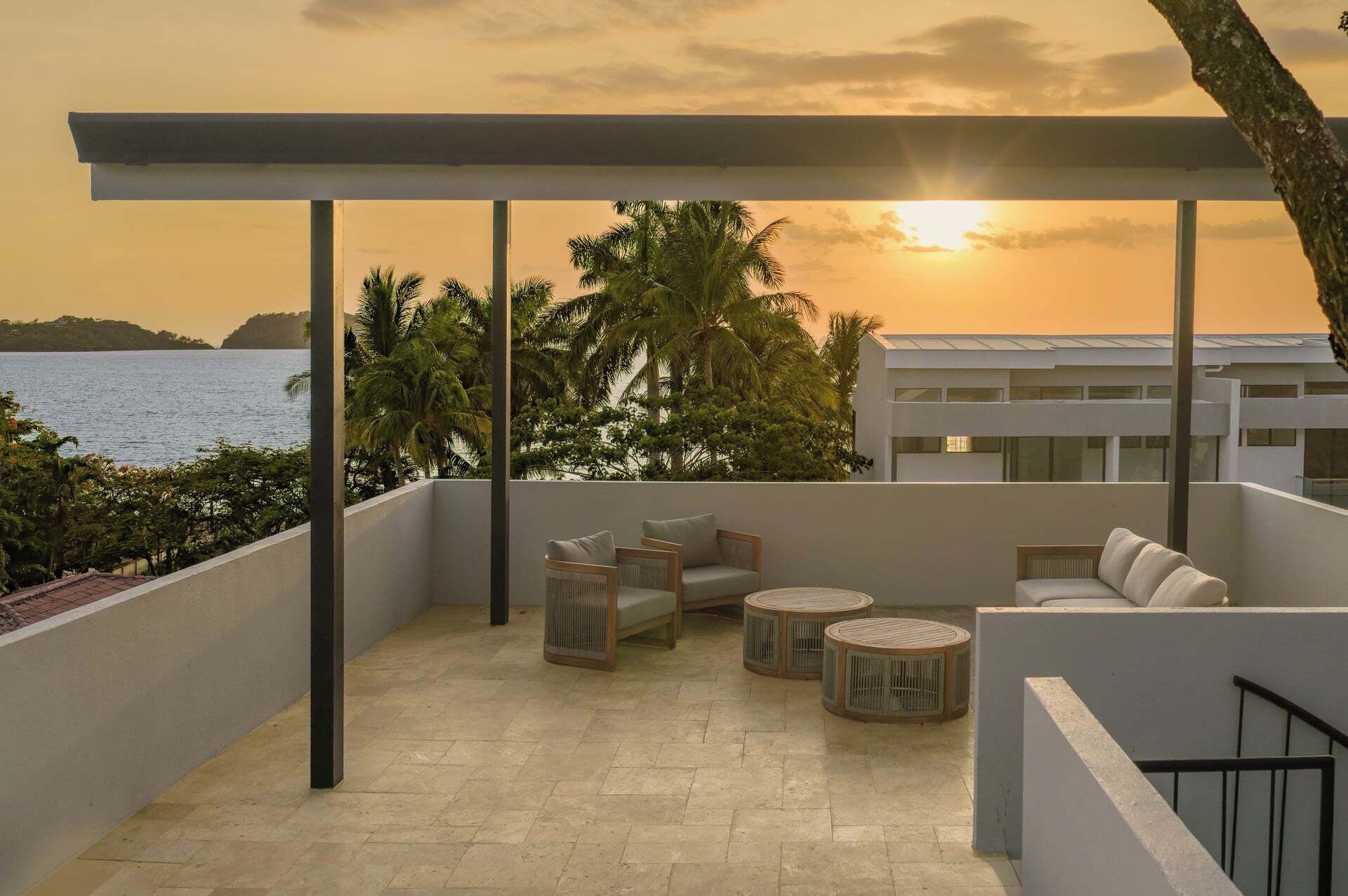 A balcony with a view of the sea and palm trees, furnished with a sofa and round tables.