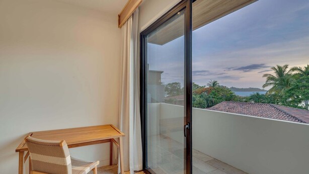 A balcony with a wooden table and chair, a sliding glass door, and a view of a coastal landscape. - Casa VistaMar Spacious 5-bedroom villa built around a tree! (Potrero)