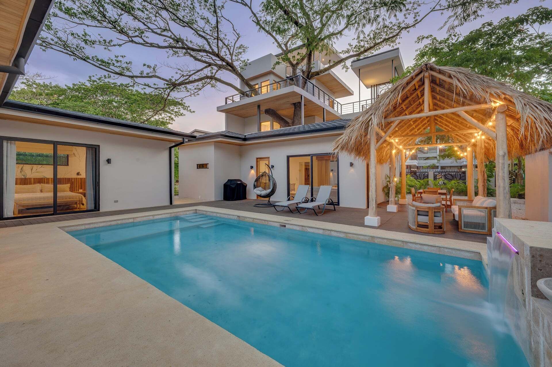 A modern poolside area with a clear pool, lounge chairs, and a thatched gazebo.
