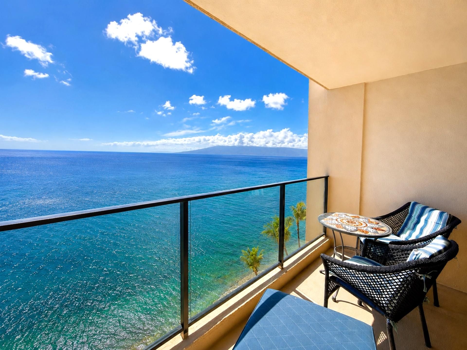 A balcony with a clear view of the ocean, a small table, two wicker chairs, and a single palm tree.