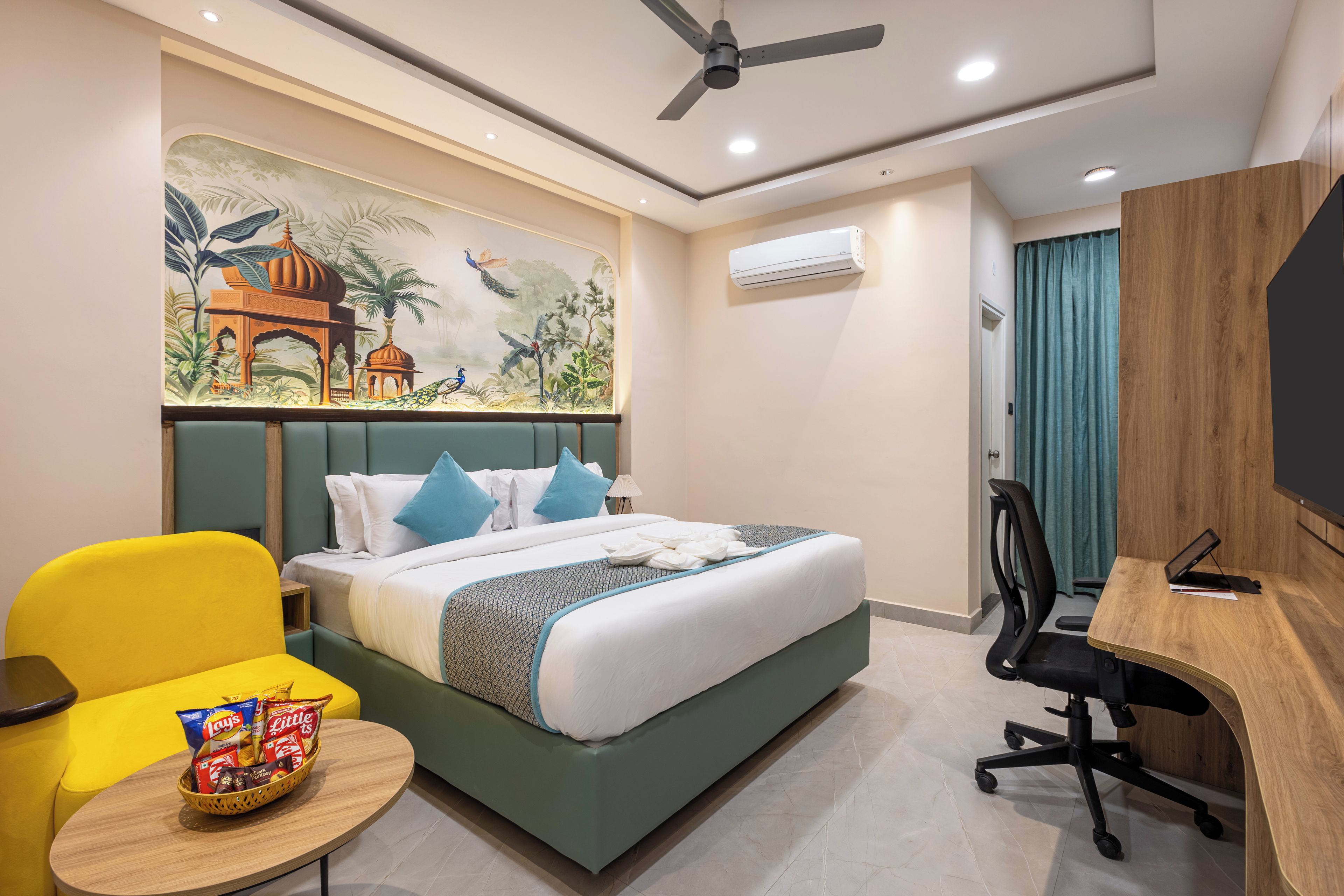 A hotel room with a bed, a desk, a chair, a wall-mounted air conditioner, and a ceiling fan.