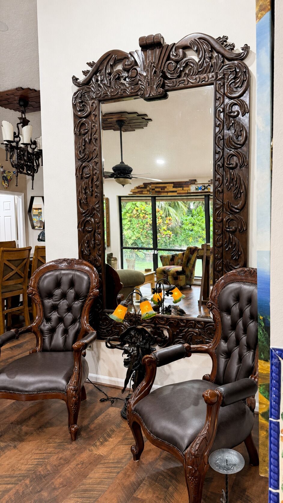 A room with a large ornate mirror, two upholstered chairs, and a window with a view of greenery.