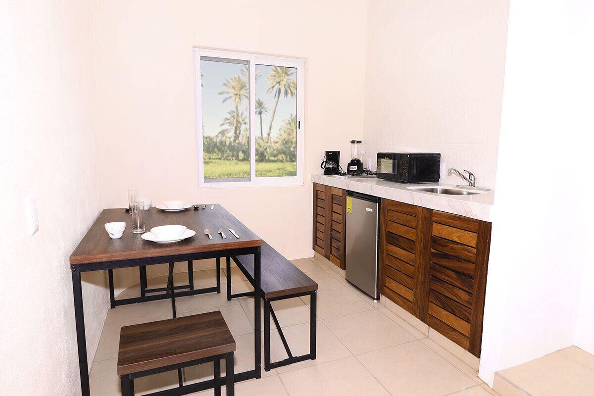 Cocina compacta con mesa de comedor, banco y vista a palmeras a través de una ventana.