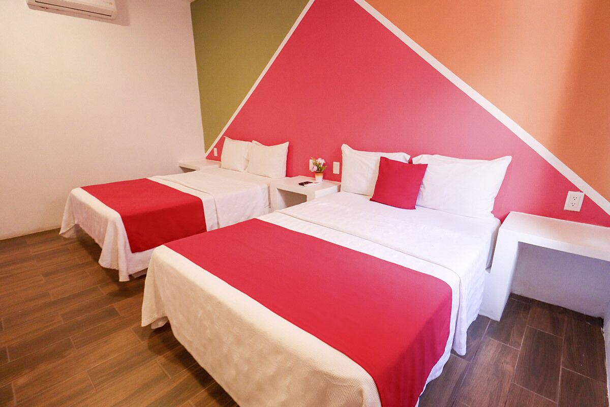Habitación de hotel con dos camas, cada una con una colcha rojo y blanco, una mesita de noche con lámpara y un equipo de aire acondicionado instalado en la pared.