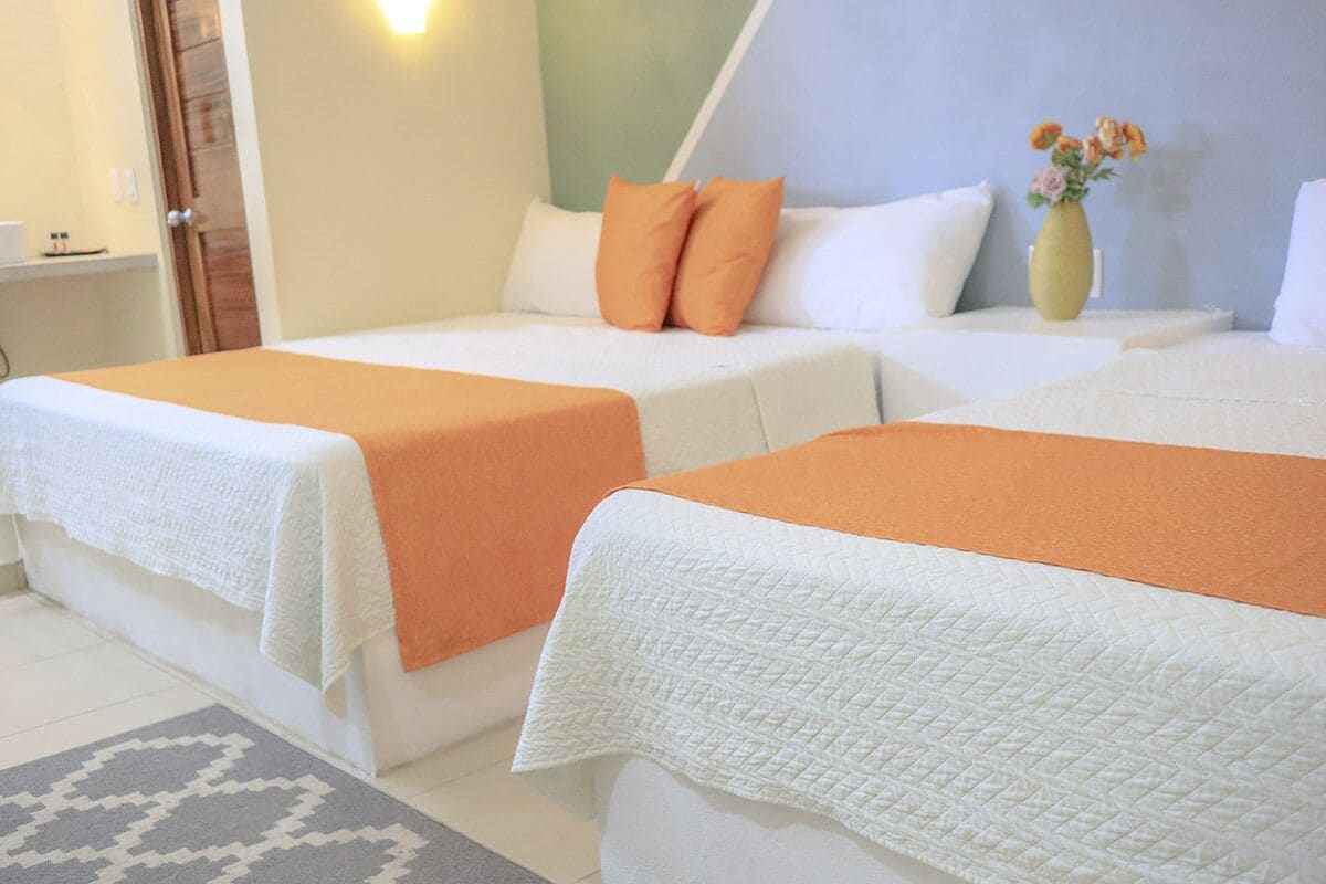 Une chambre d’hôtel avec deux lits, chacun recouvert d’une couverture orange, agrémenté d’oreillers blancs et doté d’une tête de lit blanche matelassée. Un vase rempli de fleurs est posé sur la table de chevet.