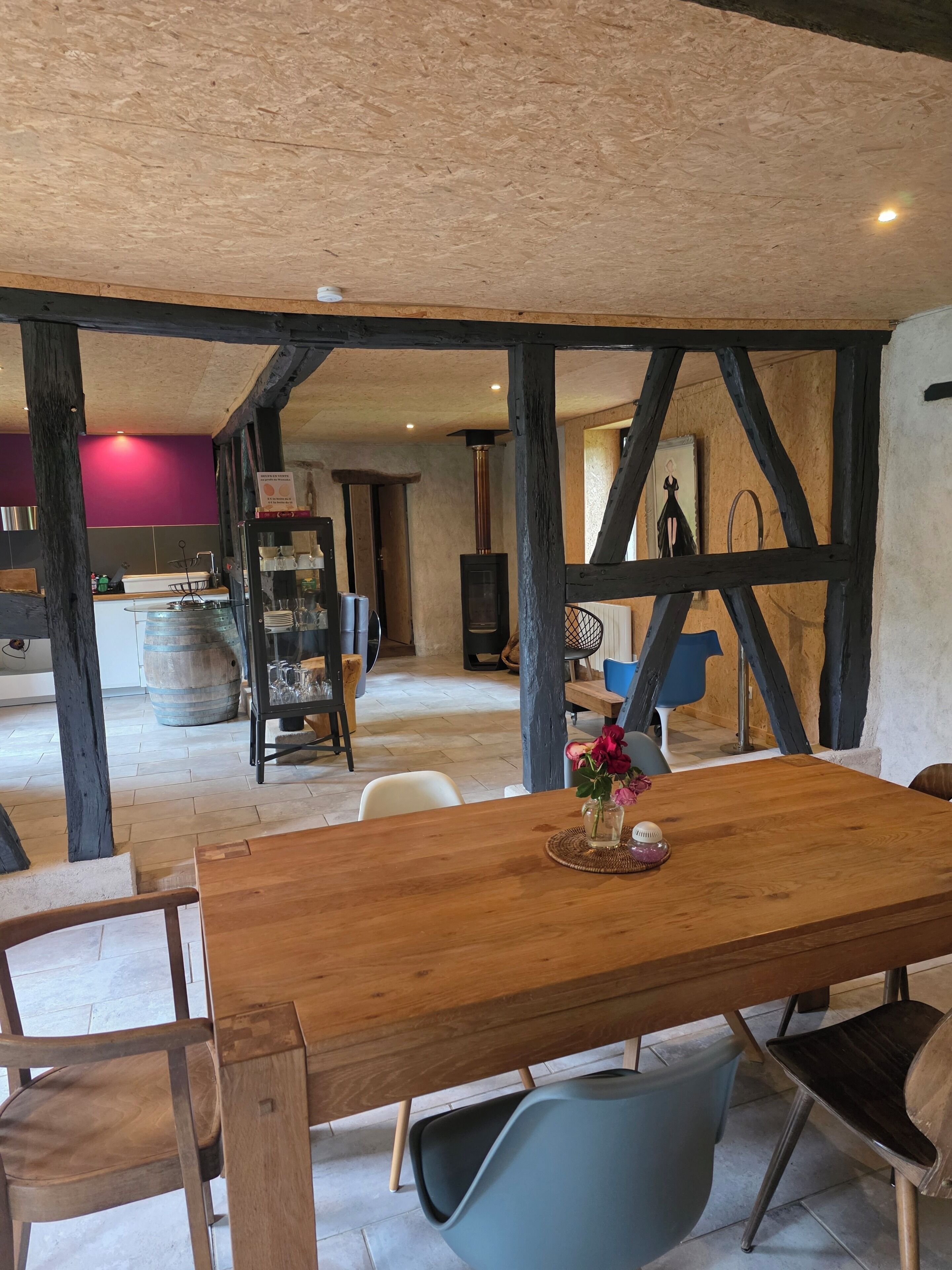 Una mesa de comedor de madera con sillas, un florero con flores y un interior rústico con vigas a la vista y una zona de cocina al fondo.