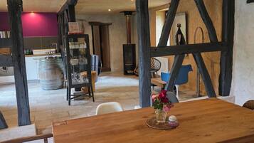 Una mesa de comedor de madera con sillas, un florero con flores y un interior rústico con vigas a la vista y una zona de cocina al fondo.