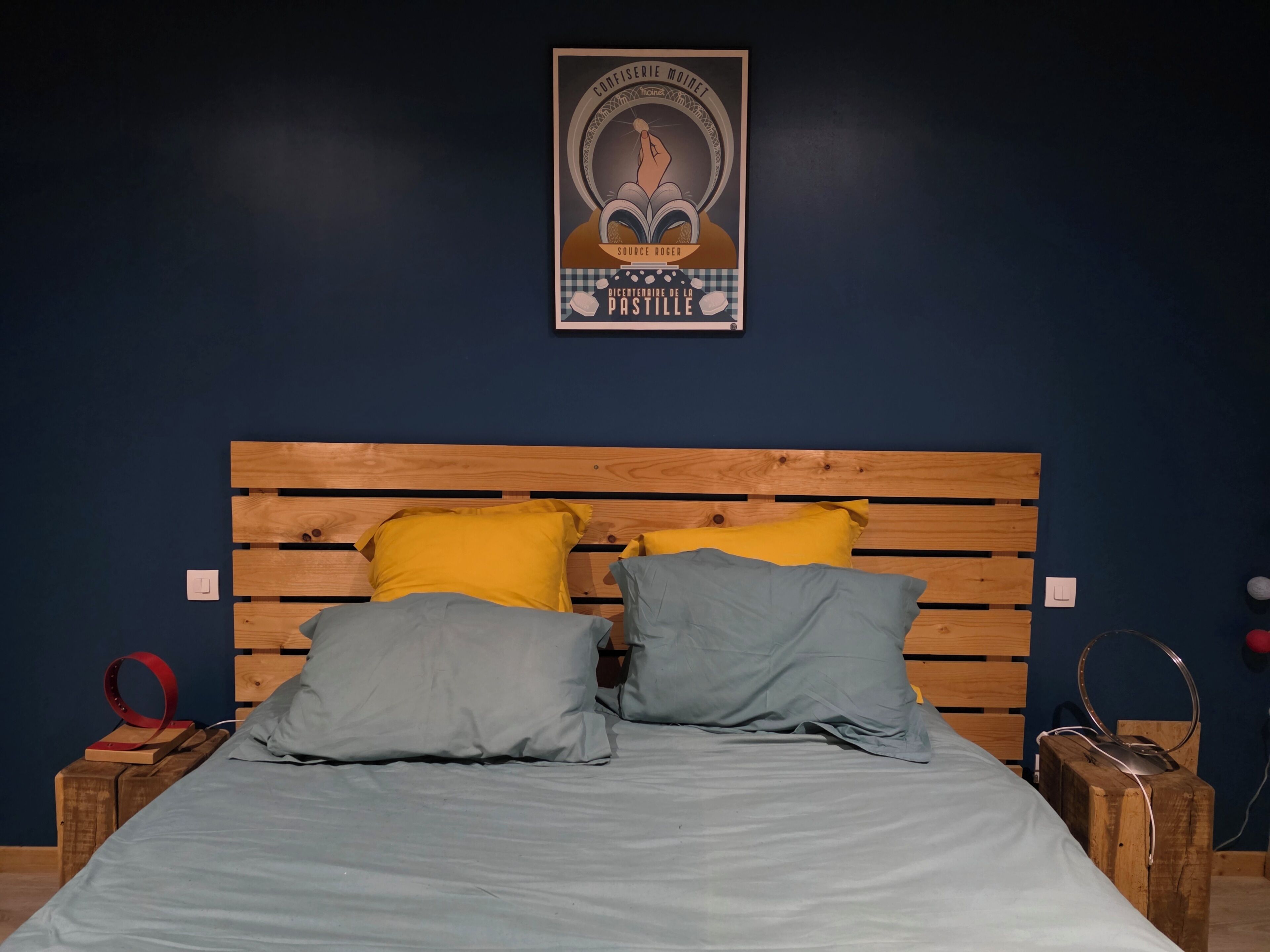A bedroom with a wooden headboard, two pillows, a bedspread, a poster on the wall, and a red circular object on a wooden box.