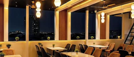 A restaurant with tables and chairs arranged for dining, large windows with views of the cityscape, and ceiling fans.