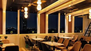 A restaurant with tables and chairs arranged for dining, large windows with views of the cityscape, and ceiling fans. - Erleen Hotel & Rooftop Restaurant (Indore)