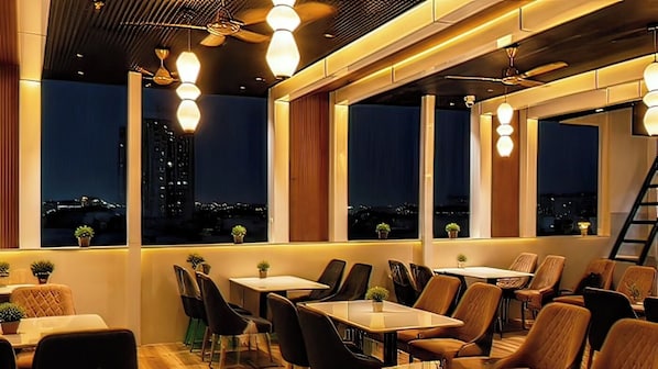 A restaurant with tables and chairs arranged for dining, large windows with views of the cityscape, and ceiling fans. - Erleen Hotel & Rooftop Restaurant (Indore)