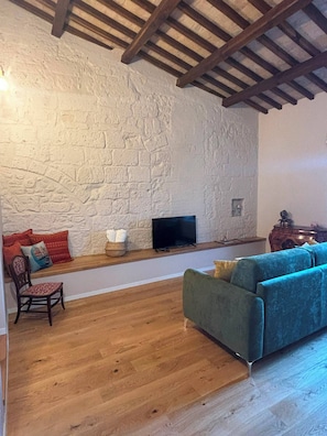 A living room with a stone wall, wooden floor, a blue sofa, a wooden side table, and a television. - Loggia Dei Mercanti 16 (Orvieto)