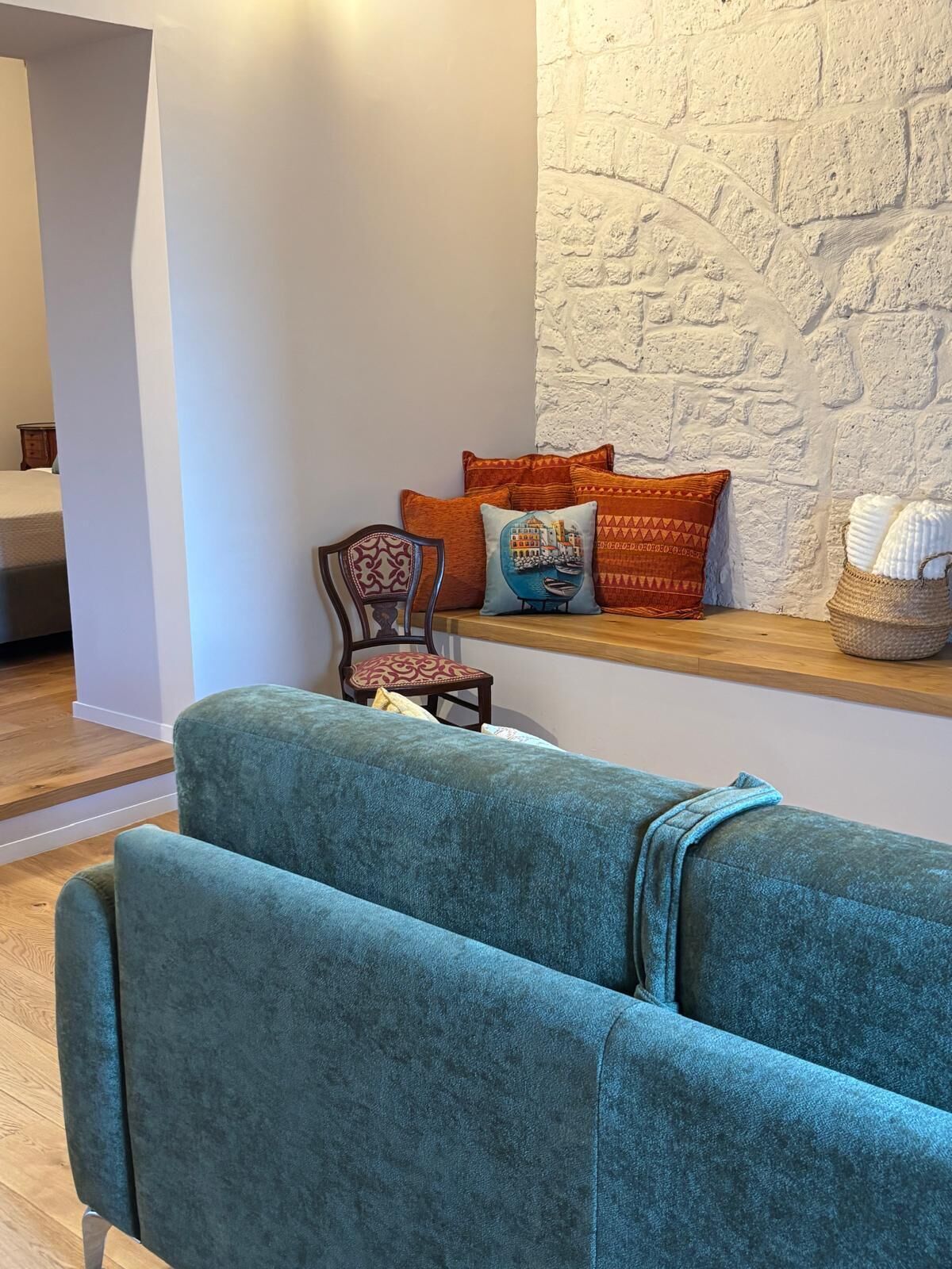 Una habitación con un sofá color turquesa, un banco de madera y una silla con cojines estampados. También se aprecia una pared blanca con textura y una estantería de madera con almohadones decorativos.