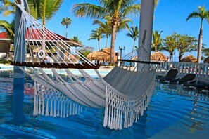 Eine Hängematte über einem Pool, im Hintergrund Palmen und ein Häuschen mit Reetdach.