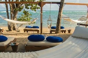 Ein Strandbereich zum Entspannen mit Hängematten und Liegestühlen, einige mit blauen Kissen, und einer weißen Sonnenschutzkonstruktion.