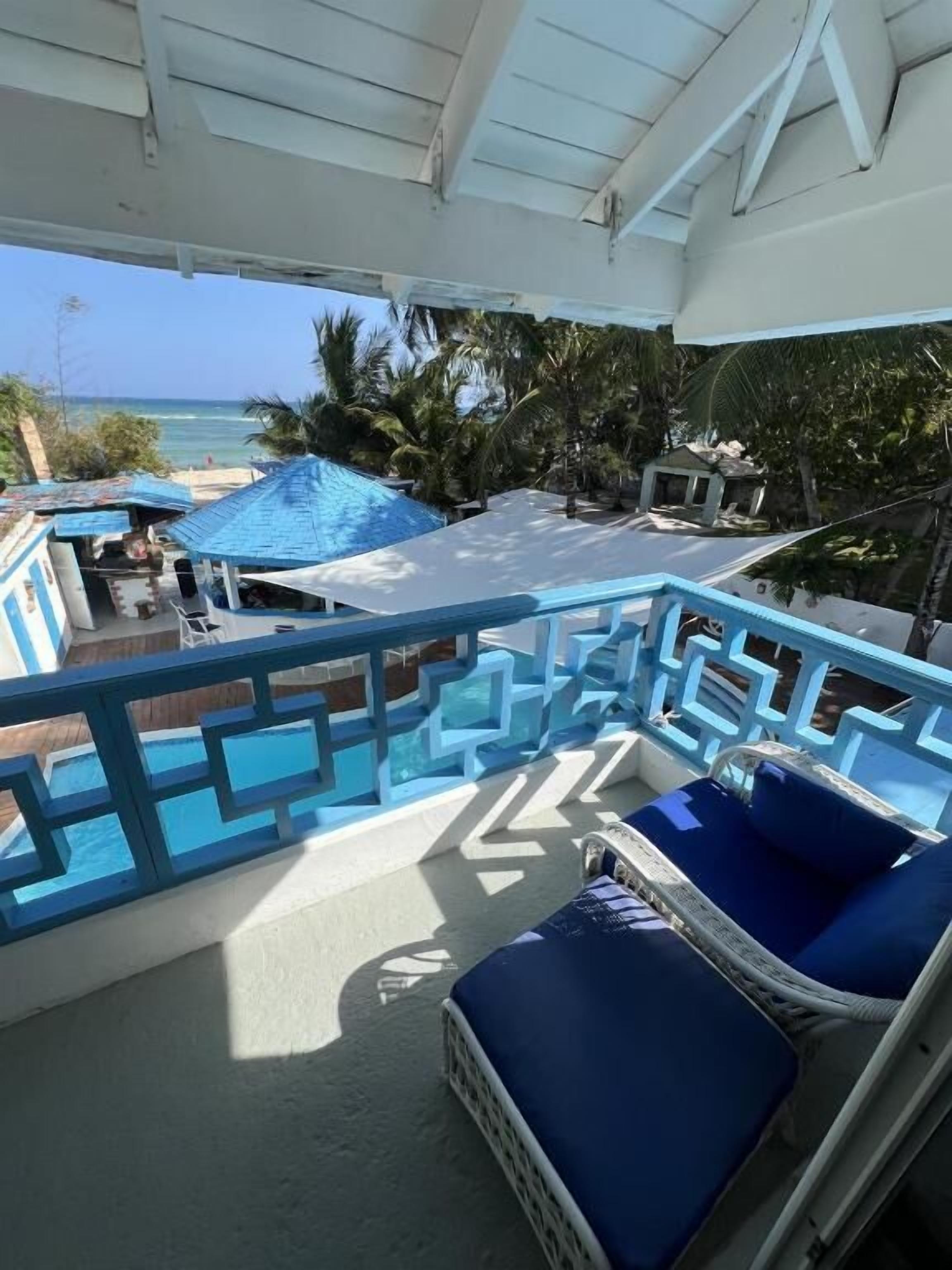A balcony with a view of the ocean, a pool area, and a lounge chair.