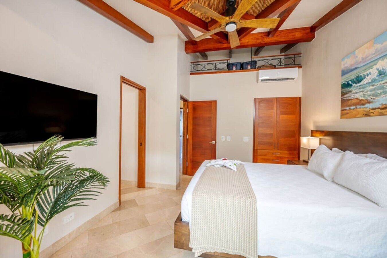 A hotel room with a bed, a television mounted on the wall, a ceiling fan, and a painting of a beach scene.
