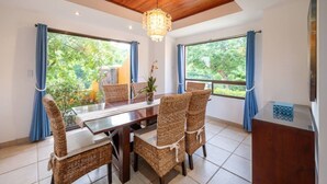 Un espace repas doté d’un plafond en bois, d’un lustre, de chaises en osier, d’une table en verre et de rideaux.