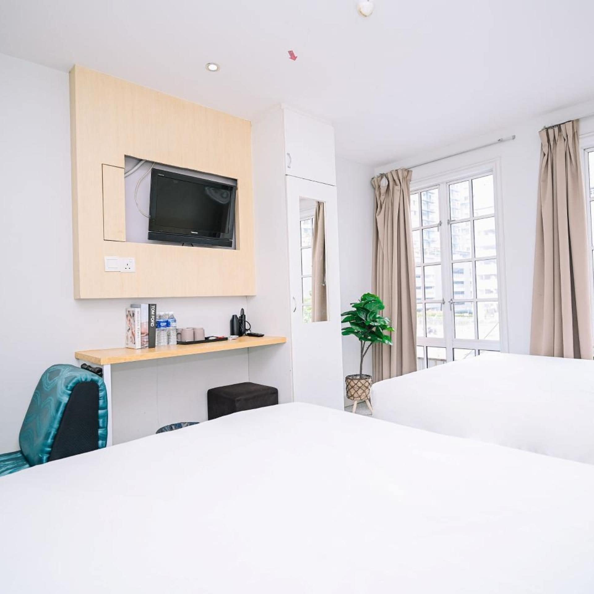 A hotel room with a bed, a desk with a chair, a television mounted on the wall, a window with curtains, and a potted plant.