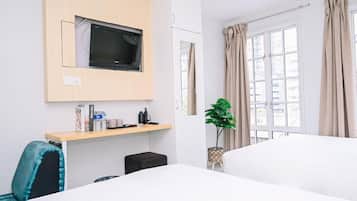 A hotel room with a bed, a desk with a chair, a television mounted on the wall, a window with curtains, and a potted plant.
