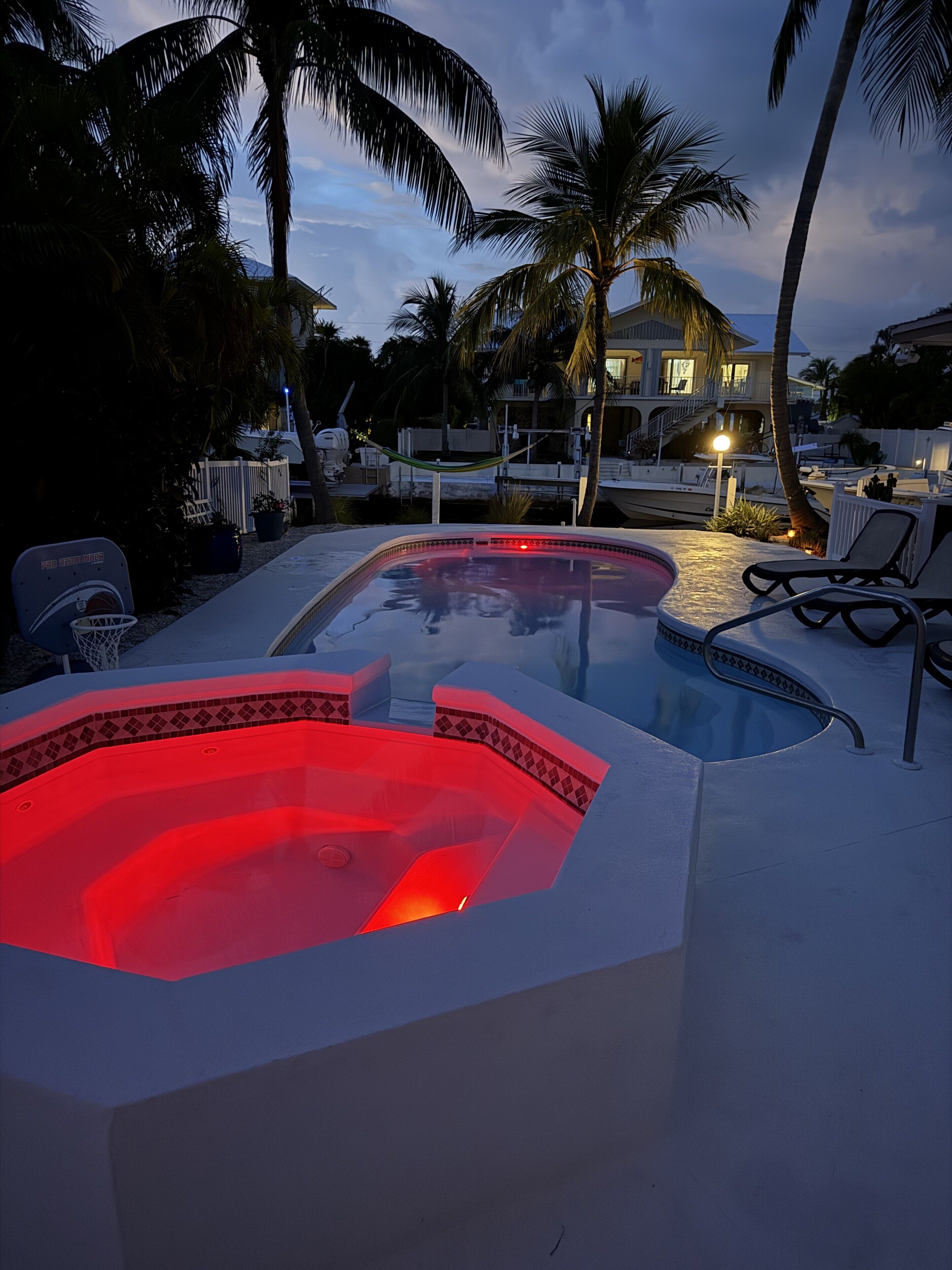 A swimming pool with a hot tub, surrounded by lounge chairs and palm trees, with a house in the background.