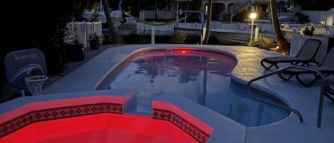 A swimming pool with a hot tub, surrounded by lounge chairs and palm trees, with a house in the background.