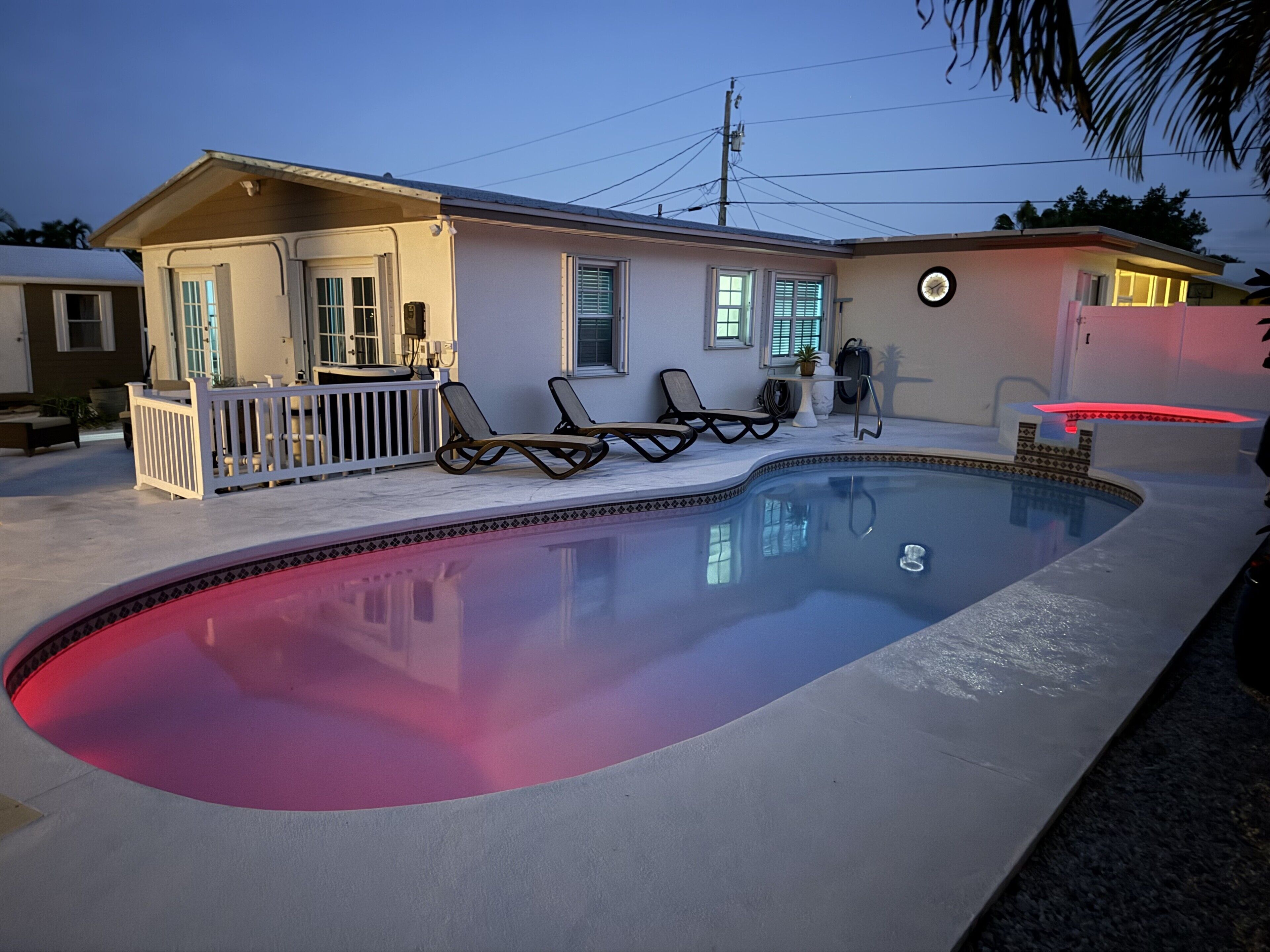 Una piscina con borde rojo, dos sillas de descanso, una pequeña terraza y una casa con porche.