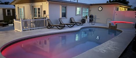 Una piscina con borde rojo, dos sillas de descanso, una pequeña terraza y una casa con porche.