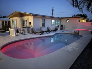A swimming pool with a red edge, two lounge chairs, a small deck, and a house with a porch.