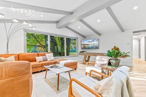 A modern living room with a sofa, coffee table, and chairs, large windows with a view of greenery, and a flat-screen TV mounted on the wall. - Park like setting on the Applegate River great for wine tasting & family reunion (Jacksonville)