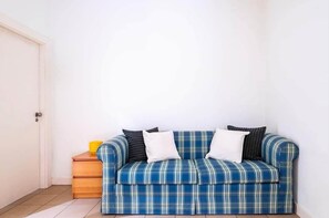 A room with a blue plaid sofa against a white wall, a wooden nightstand with a yellow object, and a closed door. - SunDream Apart I (Funchal)