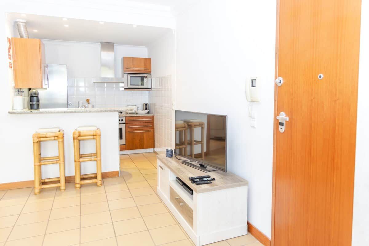 A compact kitchen with a microwave, stove, and wooden cabinets. There are two stools and a television on a stand.