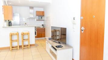 A compact kitchen with a microwave, stove, and wooden cabinets. There are two stools and a television on a stand.