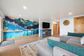 Ein Wohnzimmer mit einem großen Wandgemälde, einem Flachbildfernseher, einer Holztür und einer Couch mit Kissen.