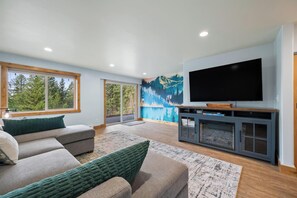 A modern living room with a large flat-screen TV, a sectional sofa, and a mural of a forest. - Minutes to Whitefish Lake and Mountain (Whitefish)