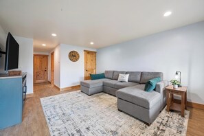 Ein modernes Wohnzimmer mit einem grauen Ecksofa, einer Holztür, einer Wanduhr und einem Fernseher auf einem Schrank.
