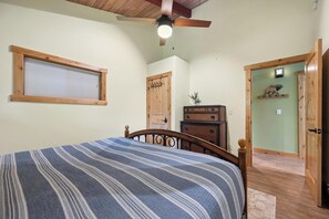Ein Schlafzimmer mit einem Bett, einer Holzkommode, einem Deckenventilator, einem Fenster mit Jalousien und einer Tür zu einem weiteren Raum.