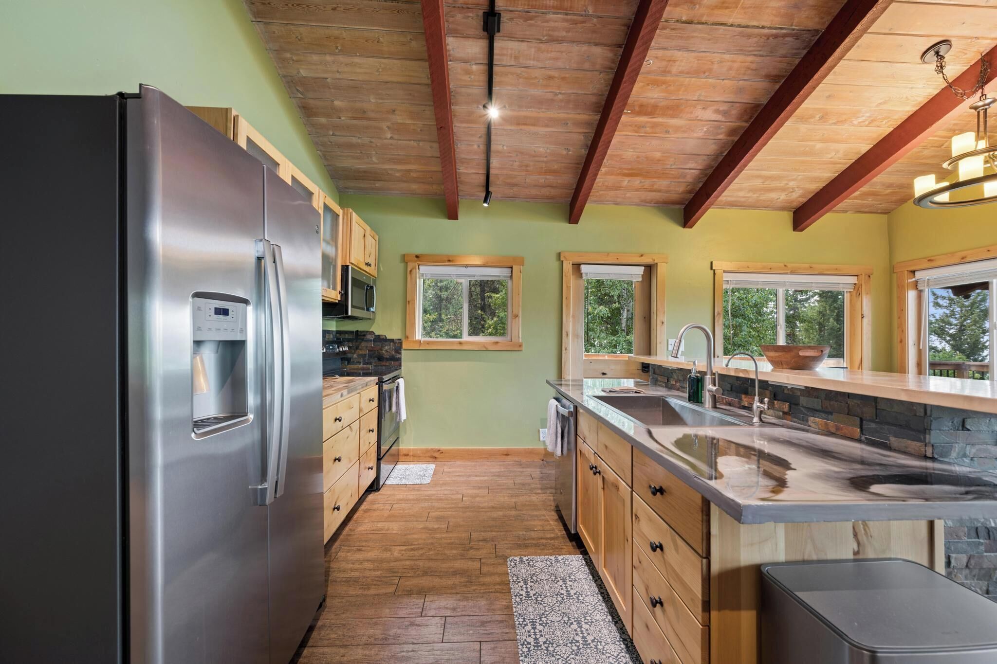 A kitchen with a stainless steel refrigerator, wooden cabinets, a stone countertop, and a wooden ceiling.